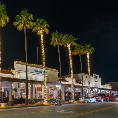 Palm trees wrapped in lights line a lively downtown street in Chandler, Arizona, with brightly lit storefronts and parked cars at night.