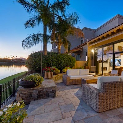 A cozy outdoor patio with wicker furniture overlooks a calm lake at sunset, framed by lush palm trees and soft outdoor lighting for a relaxing ambiance.