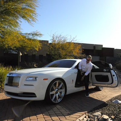 A man steps out of a white Rolls-Royce parked on a driveway, with a sports car visible in the background.
