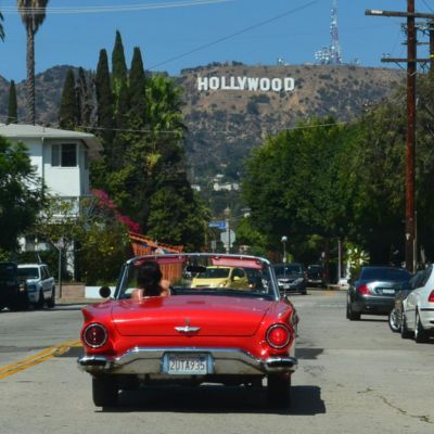 A classic red convertible driving toward the Hollywood sign on a sunny street in Los Angeles.