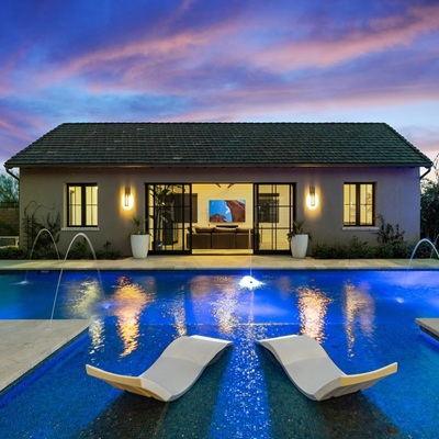 Backyard pool with water features, lounge chairs in the water, and a guest house illuminated at twilight.