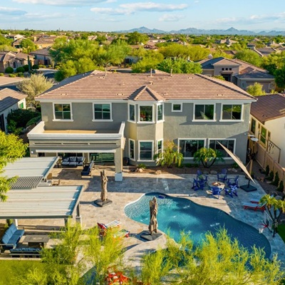 An aerial view of a spacious suburban home featuring a large backyard with a swimming pool, shaded patio areas, and landscaped surroundings in a quiet neighborhood.