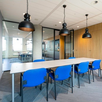 A small conference room with a long white table, blue chairs, and black pendant lighting, surrounded by wood-paneled walls and glass partitions.