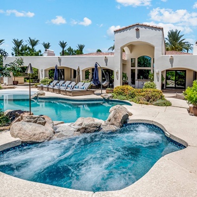 Resort-style pool and spa with lounge chairs, surrounded by lush landscaping and a beautiful white stucco home.