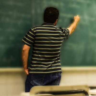 A teacher writing on a chalkboard in an empty classroom, viewed from behind.
