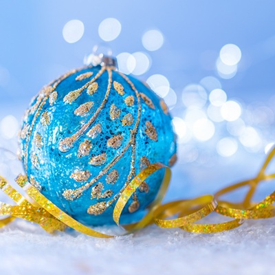 Blue ornament with gold details resting on snow, surrounded by golden ribbon and sparkling lights in the background.