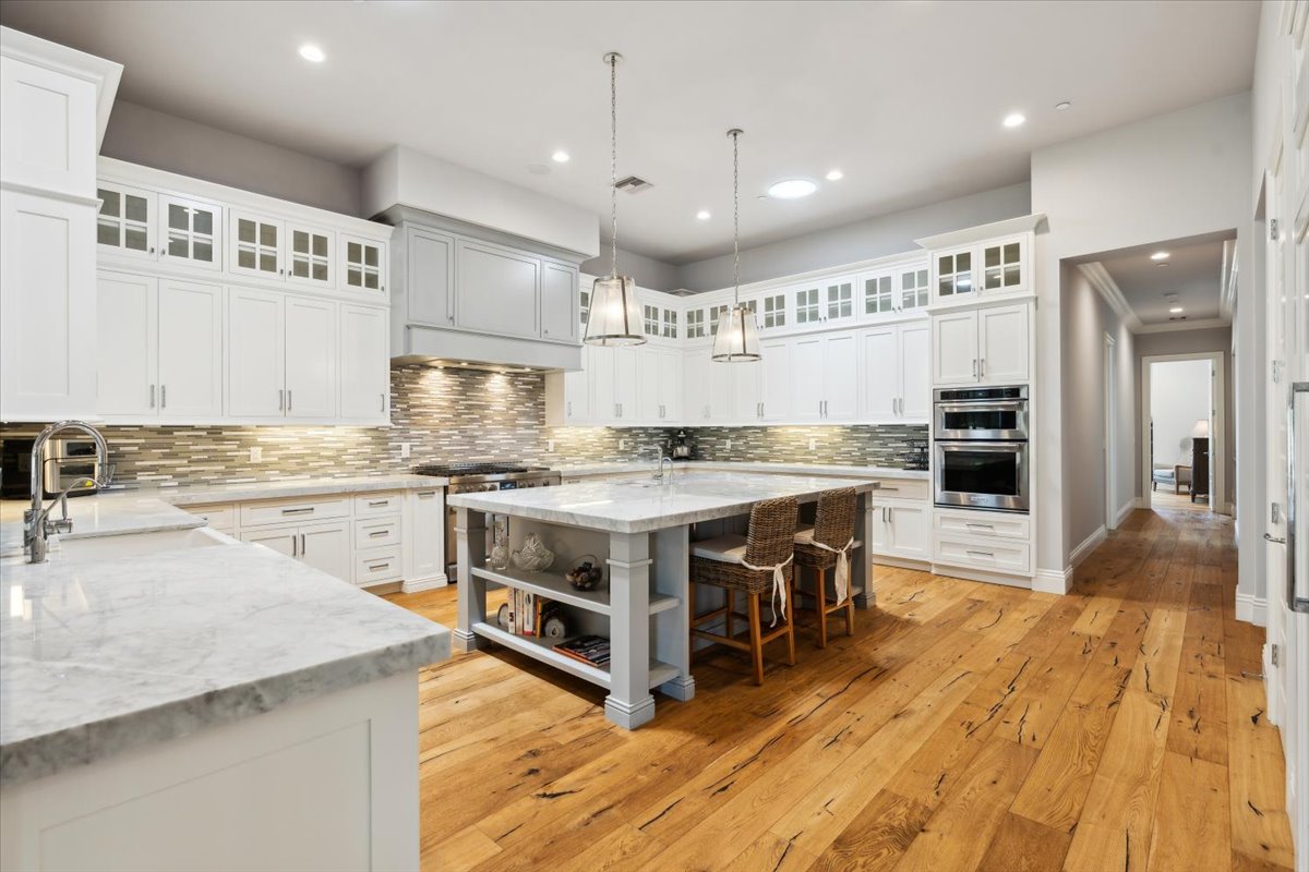 A spacious kitchen with white cabinetry, a large marble island with seating, wood flooring, and pendant lighting. The backsplash features a modern tile design.