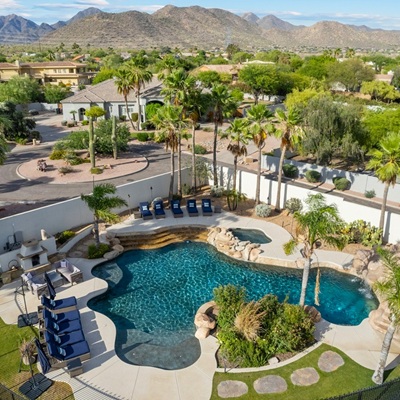 Aerial view of a backyard in a suburban neighborhood featuring a resort-style pool with a waterfall, lush landscaping, palm trees, and a mountain backdrop.
