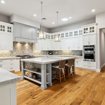 A spacious kitchen with white cabinetry, a large marble island with seating, wood flooring, and pendant lighting. The backsplash features a modern tile design.