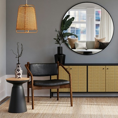 A modern living room corner with a round mirror, woven cabinet, black chair, potted plants, and a hanging rattan light fixture.