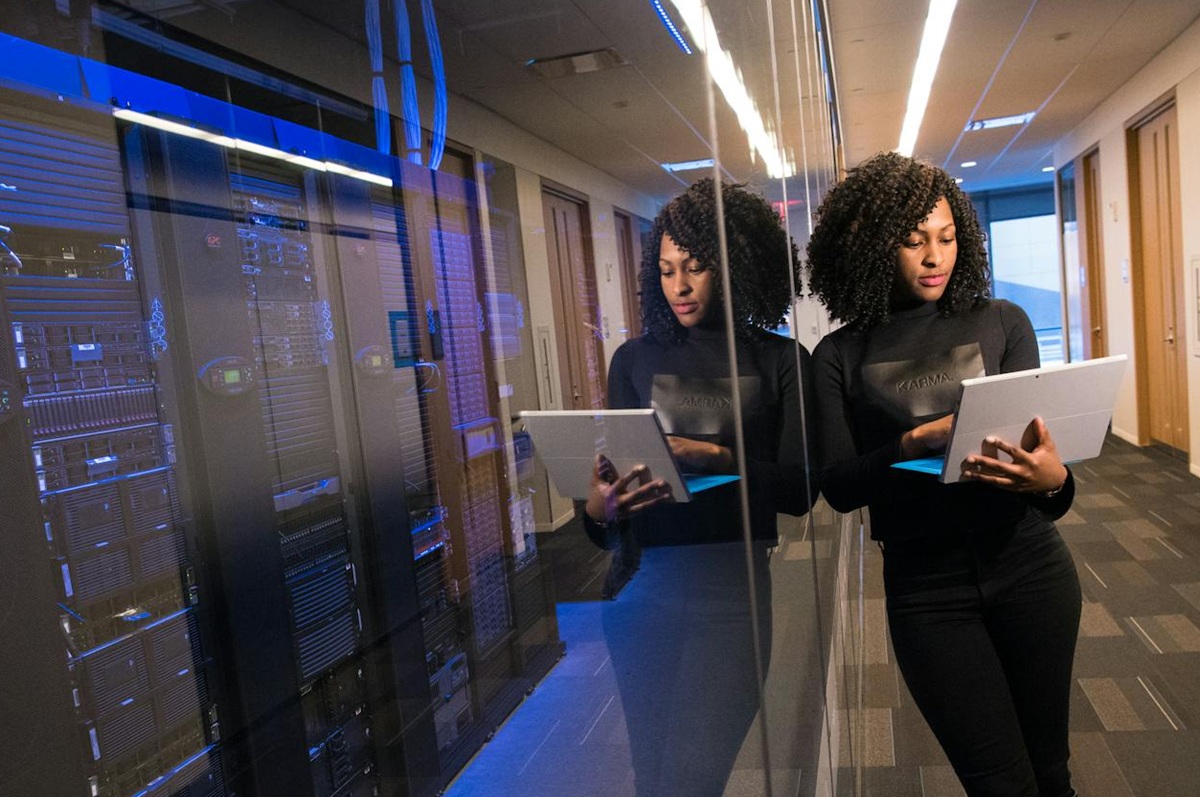 An African American woman  stands in a server room, using a tablet while her reflection appears on the glass.