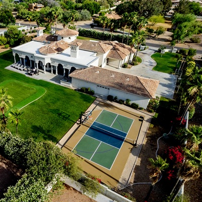 An aerial view of a Mediterranean-style estate with a lush lawn, a pool, a pickleball court, and palm trees surrounding the property.
