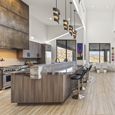 A modern open-concept kitchen with a sleek island, bar seating, pendant lighting, and high ceilings. The space features warm wood tones, metallic finishes, and large windows revealing scenic desert views.