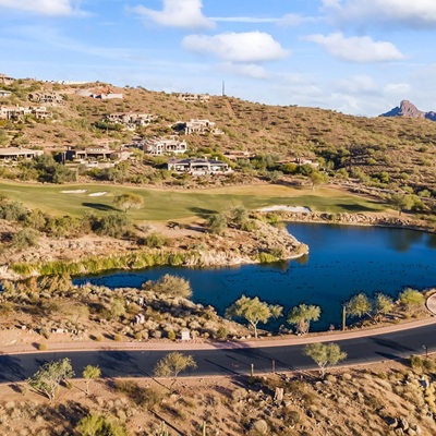 A scenic desert golf course community with luxury homes, a lake, and mountain views under a bright blue sky.