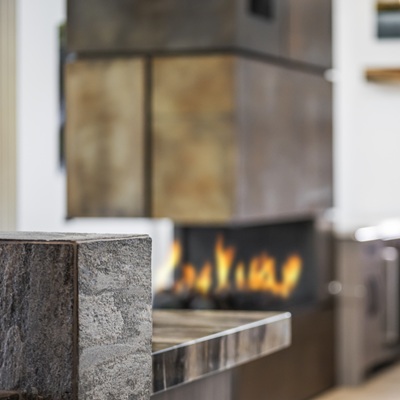 A modern kitchen with a stone countertop in the foreground and a sleek, double-sided fireplace with flames visible in the middle. The background features contemporary cabinetry and shelving.