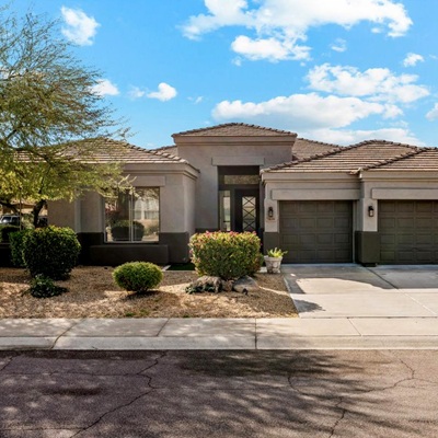 Single-story home with a three-car garage, flat rooflines, desert landscaping, and a stucco exterior in neutral tones under a blue sky.