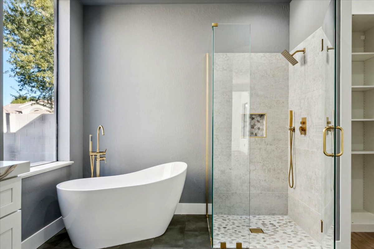 Luxury bathroom with a freestanding white soaking tub, glass-enclosed shower with gold fixtures, and a large window providing natural light.