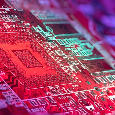 A colorful red circuit board illuminated by pink and blue lighting, showcasing fine electronic traces and component slots.