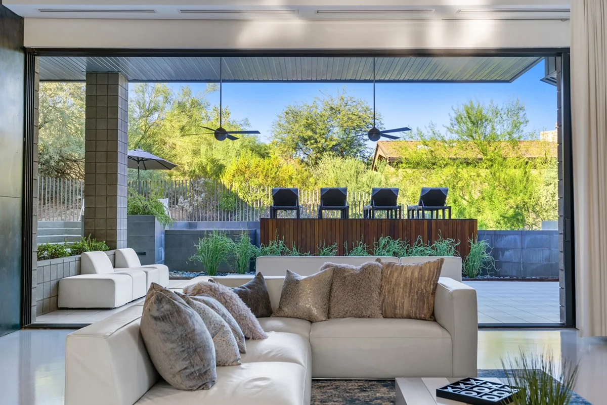 View from indoor living space looking out to a shaded patio with ceiling fans, modern outdoor furniture, and lush greenery behind a wooden privacy wall.