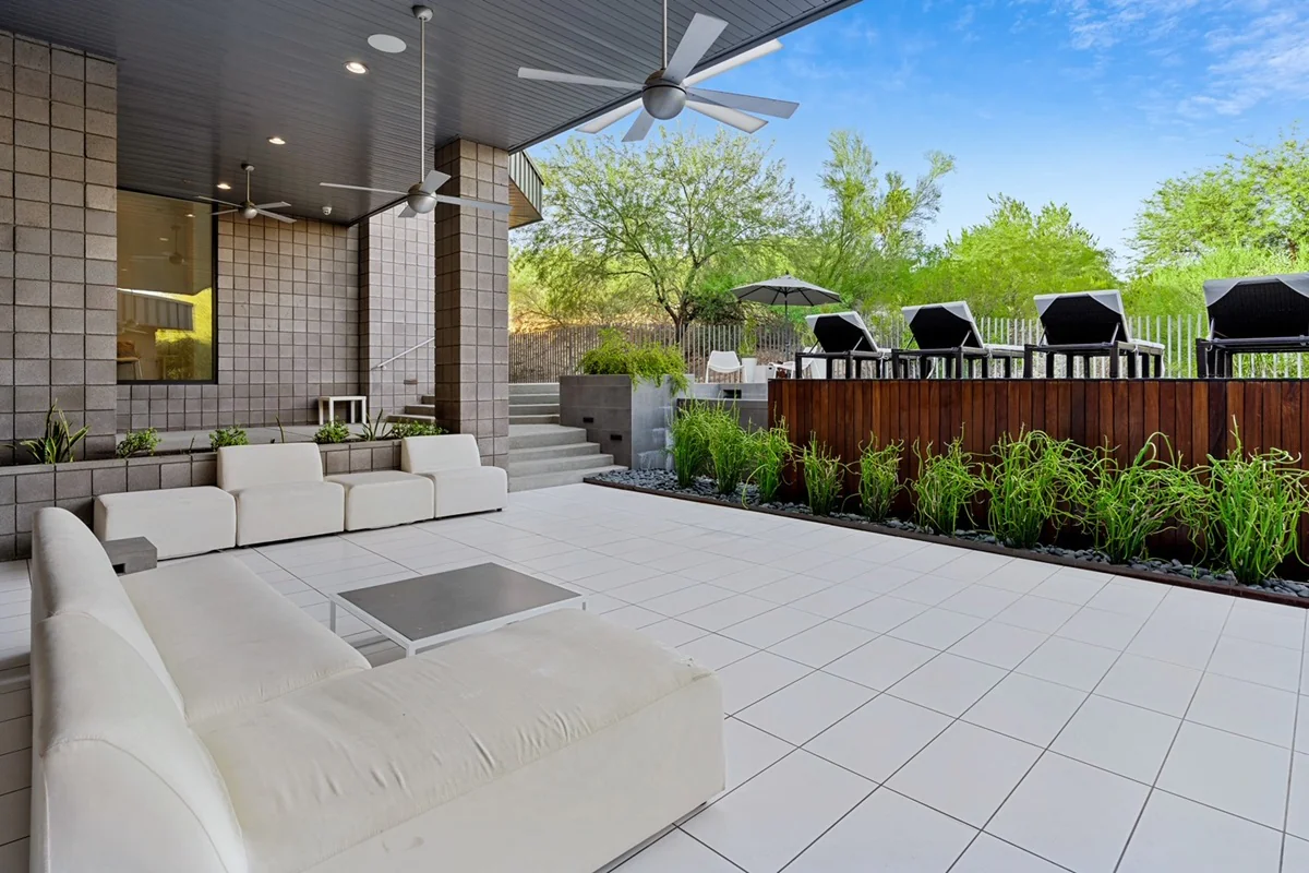 Covered outdoor patio with white modern seating, tile flooring, ceiling fans, and a raised wood-deck bar area lined with black lounge chairs and green landscaping.