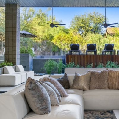 View from indoor living space looking out to a shaded patio with ceiling fans, modern outdoor furniture, and lush greenery behind a wooden privacy wall.