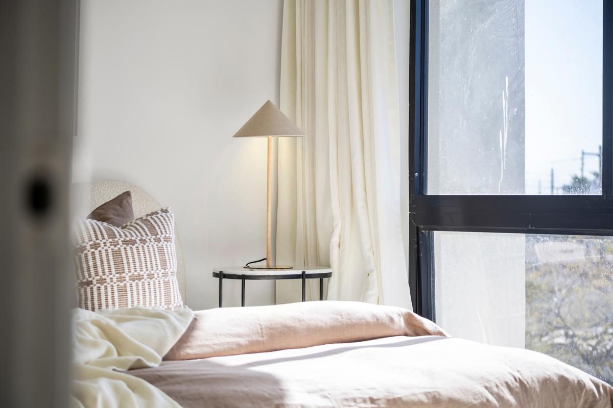 Softly lit modern bedroom with neutral tones, a tall lamp, and cozy layered bedding beside a large window.