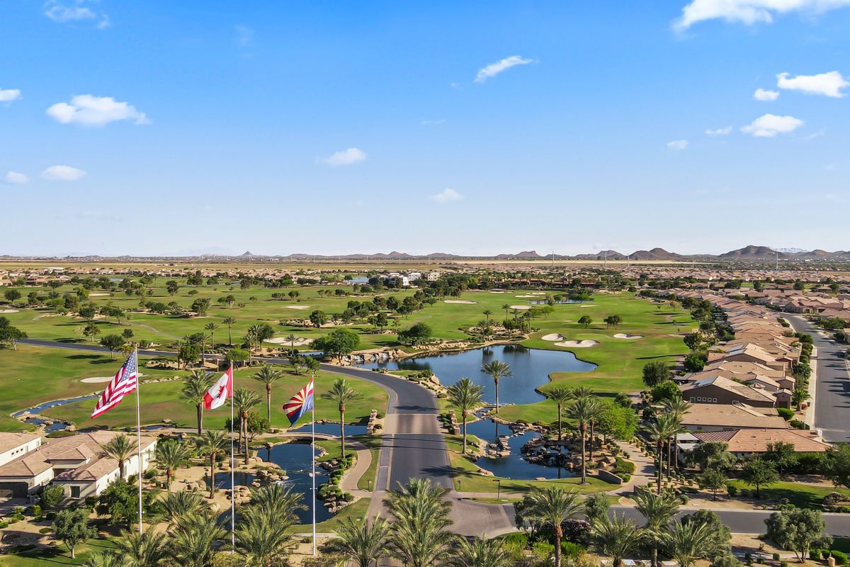 Community entrance with palm trees, flags, and water features leading into a lush golf course surrounded by desert homes.