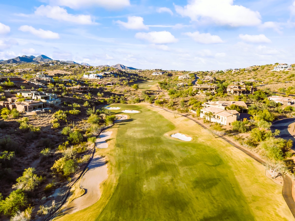 Aerial view of a green golf course lined with desert-style luxury homes and mountains in the background.